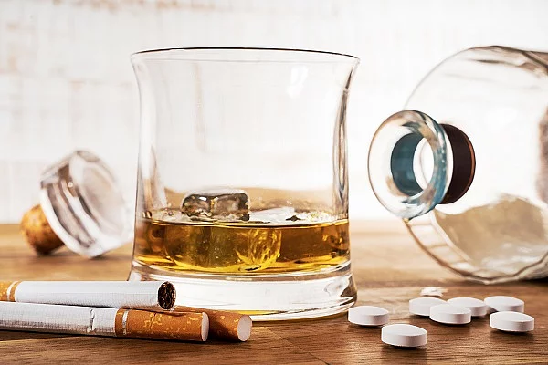 Zigaretten, Tabletten, eine liegende Glasflasche und ein Glas mit Whiskey sind auf einem Holztisch ausgebreitet.
