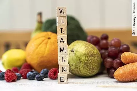 Obst und Gemüse im Hintergrund und Buchstaben auf Würfeln, die aufeinandergestapelt das Wort "Vitamine" ergeben.