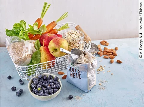 Metallkorb mit Obst, Gemüse, Haferflocken, Basilikum; daneben Mandeln und eine Schale mit Heidelbeeren