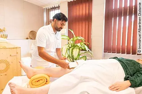 Ayurveda-Therapeut behandelt das Knie einer liegenden Patientin mit Kräuterpaste