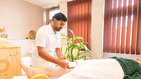 Ayurveda-Therapeut behandelt das Knie einer liegenden Patientin mit Kräuterpaste Ayurveda-Therapeut behandelt das Knie einer liegenden Patientin mit Kräuterpaste