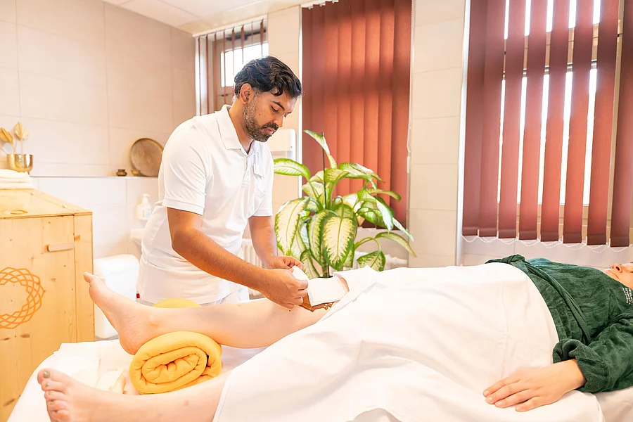 Ayurveda-Therapeut behandelt das Knie einer liegenden Patientin mit Kräuterpaste Ayurveda-Therapeut behandelt das Knie einer liegenden Patientin mit Kräuterpaste