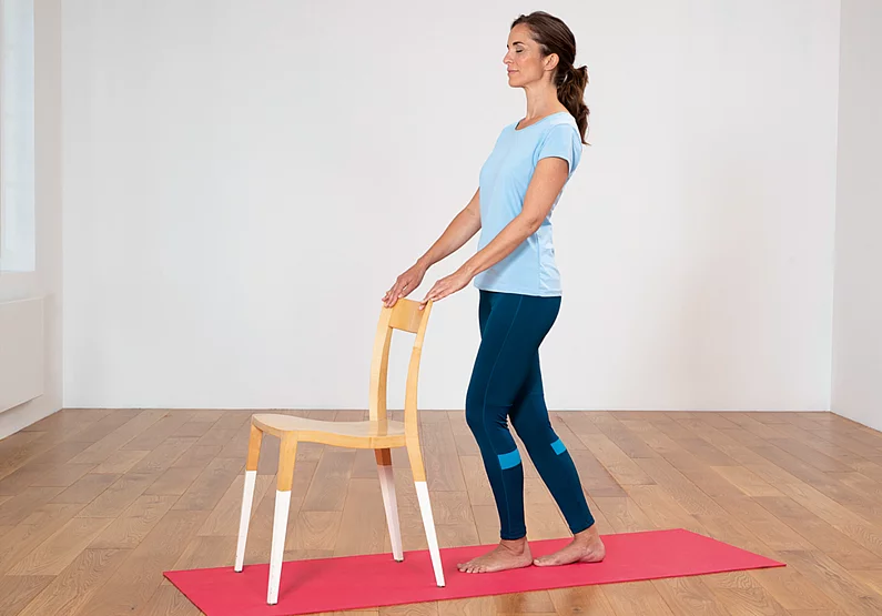 Frau steht, die Füße hintereinander, Hände auf der Lehne eines Stuhls vor ihr