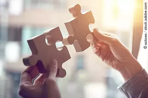 Zwei Hände, die zwei Puzzelstücke aneinander halten.