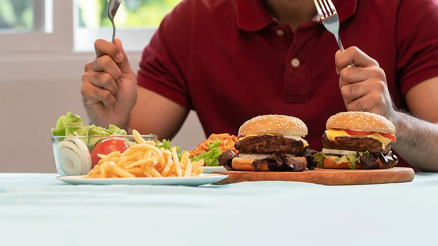 Young Man holding knife and fork are ready to eating a hamburger, French fries, and Spaghetti for Lunch. Concept of binge eating disorder (BED) and Relaxing with Eating junk food and unhealthy foods. Mann sitzt vor vollem Teller mit Fastfood