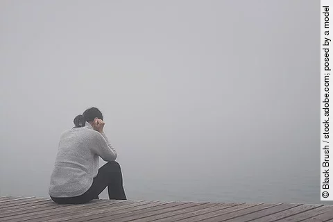 Junge Frau sitzt gekrümmt an einem Steg und vergräbt ihr Gesicht hinter ihren Händen.