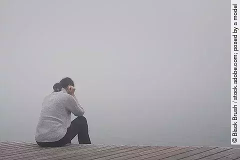 Junge Frau sitzt gekrümmt an einem Steg und vergräbt ihr Gesicht hinter ihren Händen.