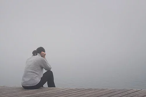 Junge Frau sitzt gekrümmt an einem Steg und vergräbt ihr Gesicht hinter ihren Händen.