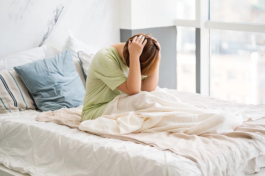 Junge Frau sitzt mit Kopfschmerzen im Bett, hält sich mit beiden Händen den schmerzenden Kopf