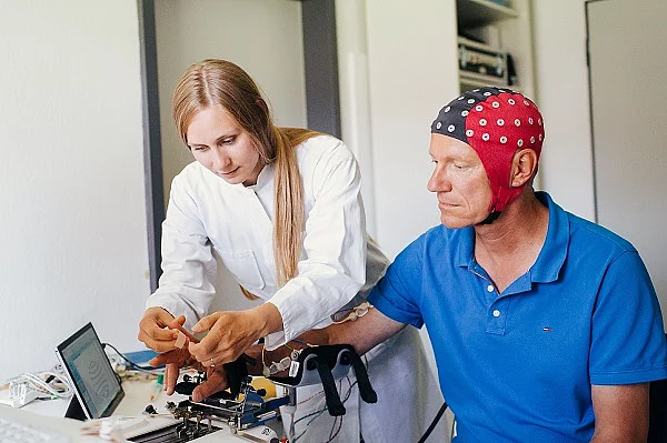 Eine Frau im Arztkittel schaut auf einen Monitor auf einem Tisch und neben ihr sitzt ein Mann, der die Neuroprothese trägt. 