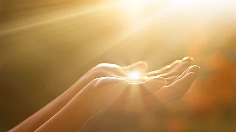 Eine Person hält die Hände wie eine Schale in die Luft. Im Hintergrund und in den Händen reflektieren Sonnenstrahlen.