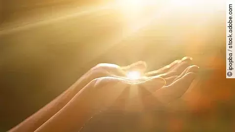 Eine Person hält die Hände wie eine Schale in die Luft. Im Hintergrund und in den Händen reflektieren Sonnenstrahlen.