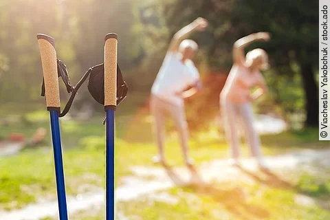 Ein älteres Ehepaar macht Sportübungen im Hintergrund (verschwommen). Im Vordergrund sind Walking Stöcke zu sehen.