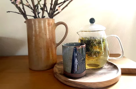 Teekanne aus Glas mit frischem Kräutertee, Teebecher aus Steingut und ein Krug mit Zweigen arrangiert auf einem Holztisch