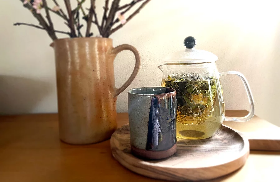 Teekanne aus Glas mit frischem Kräutertee, Teebecher aus Steingut und ein Krug mit Zweigen arrangiert auf einem Holztisch