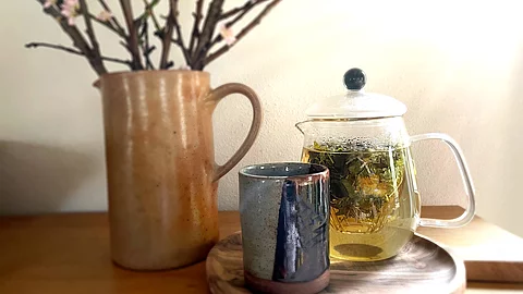 Teekanne aus Glas mit frischem Kräutertee, Teebecher aus Steingut und ein Krug mit Zweigen arrangiert auf einem Holztisch