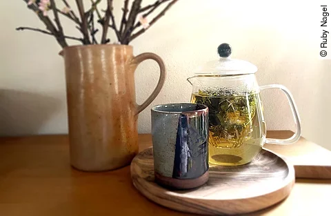 Teekanne aus Glas mit frischem Kräutertee, Teebecher aus Steingut und ein Krug mit Zweigen arrangiert auf einem Holztisch