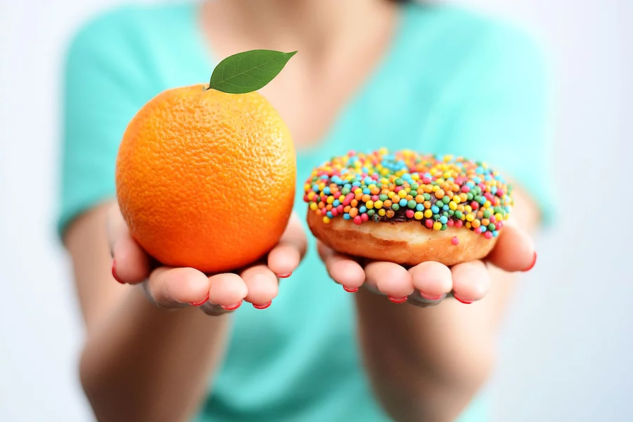 Young woman holding in hands an orange and a tasty multicolored donut, choosing healthy food or dessert Junge Frau hält eine Orange und einen Donut in die Kamera