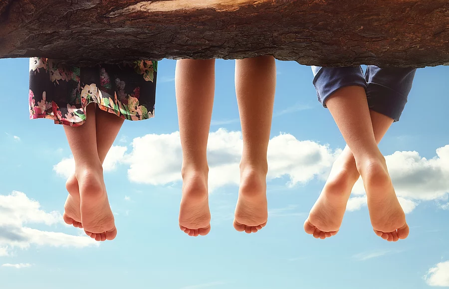 3 Kinder sitzen auf einem Baumstamm und lassen die Beine baumeln.