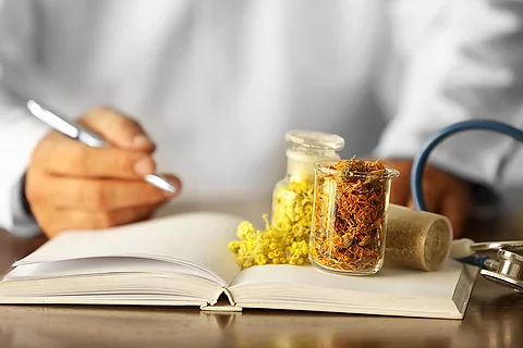 Mänlicher Arzt mit einem Rezeptbuch und Gläsern mit Naturheilpflanzen auf dem Tisch.
