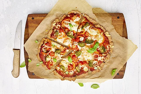 Pizza mit Tomaten, Broccoli und Käse auf einem Holzbrett, daneben liegt ein Pizzamesser
