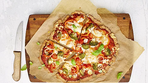 Pizza mit Tomaten, Broccoli und Käse auf einem Holzbrett, daneben liegt ein Pizzamesser