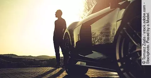 Mann steht vor seinem Sportwagen und schaut in den Sonnenuntergang