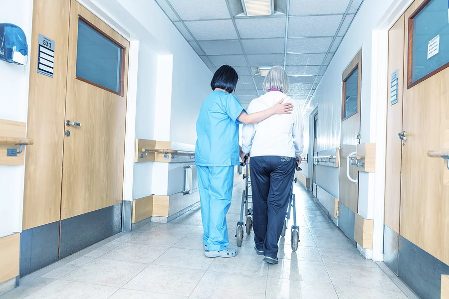 Pflegerin unterstützt Patientin beim Laufen mit ihrer Gehilfe.