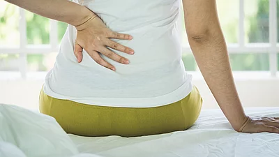 Frau auf Bettkante hält sich den schmerzenden unteren Rücken