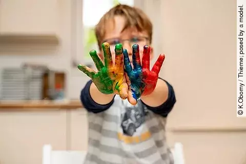 Kind mit Brille streckt die Arme nach vorne. Die Handflächen sind bunt bemalt. 