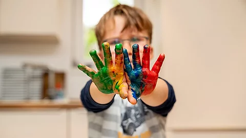 Kind mit Brille streckt die Arme nach vorne. Die Handflächen sind bunt bemalt.