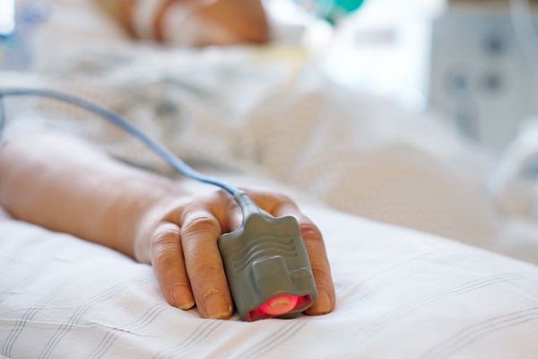 Patient liegt in seinem Bett auf der Intensivstation. Person ist nicht erkennbar, nur die Hand mit einem Finger im Pulsoximeter. 