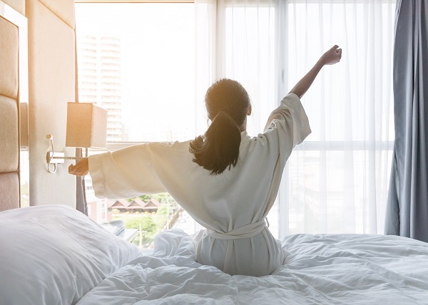 Frau sitzt auf dem Bett und streckt die Arme zur Seite. 